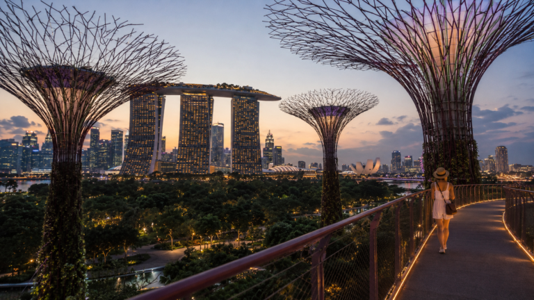 إطلالة بانورامية على حدائق الخليج في سنغافورة وقت الغروب، مع أشجار Supertree Grove وفندق Marina Bay Sands ومسافرة تسير على الممشى المرتفع.